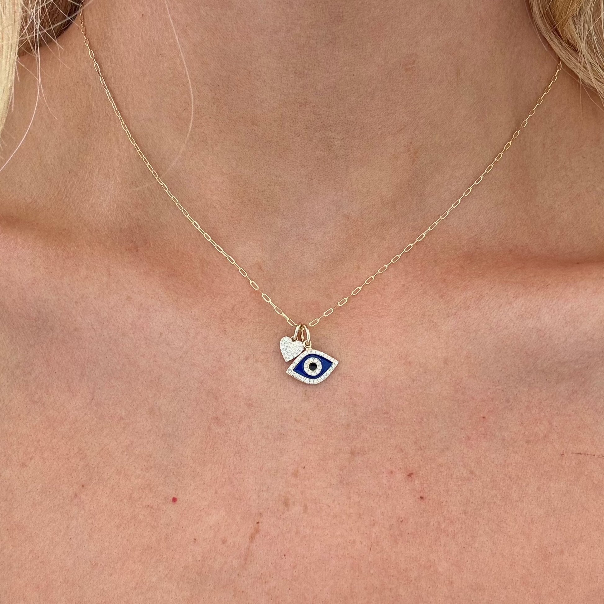 A close-up image of a person's neckline adorned with the TALIA Charm Necklace by Dylan James Jewelry. The 14kt yellow gold chain features a pendant with an eye design in blue and white hues, complemented by a small diamond heart.