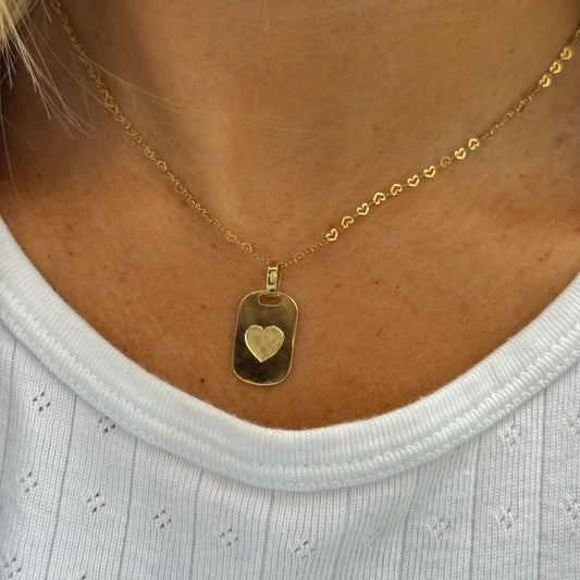 The LIBBY Necklace by Dylan James Jewelry features a hand-engraved yellow gold heart pendant on a heart chain with delicate heart-shaped links, perfect for wearing elegantly over a light-colored shirt.