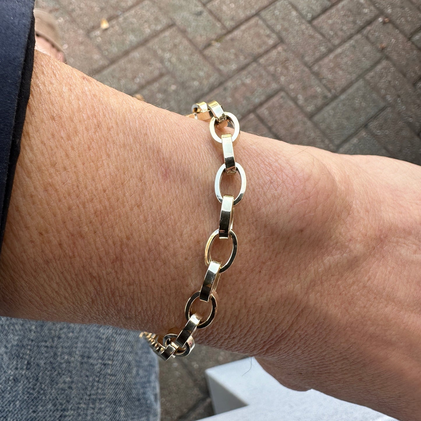 The OMAR Gold Bracelet by Dylan James Jewelry adorns a person's wrist, photographed from an overhead perspective against a brick pavement background.