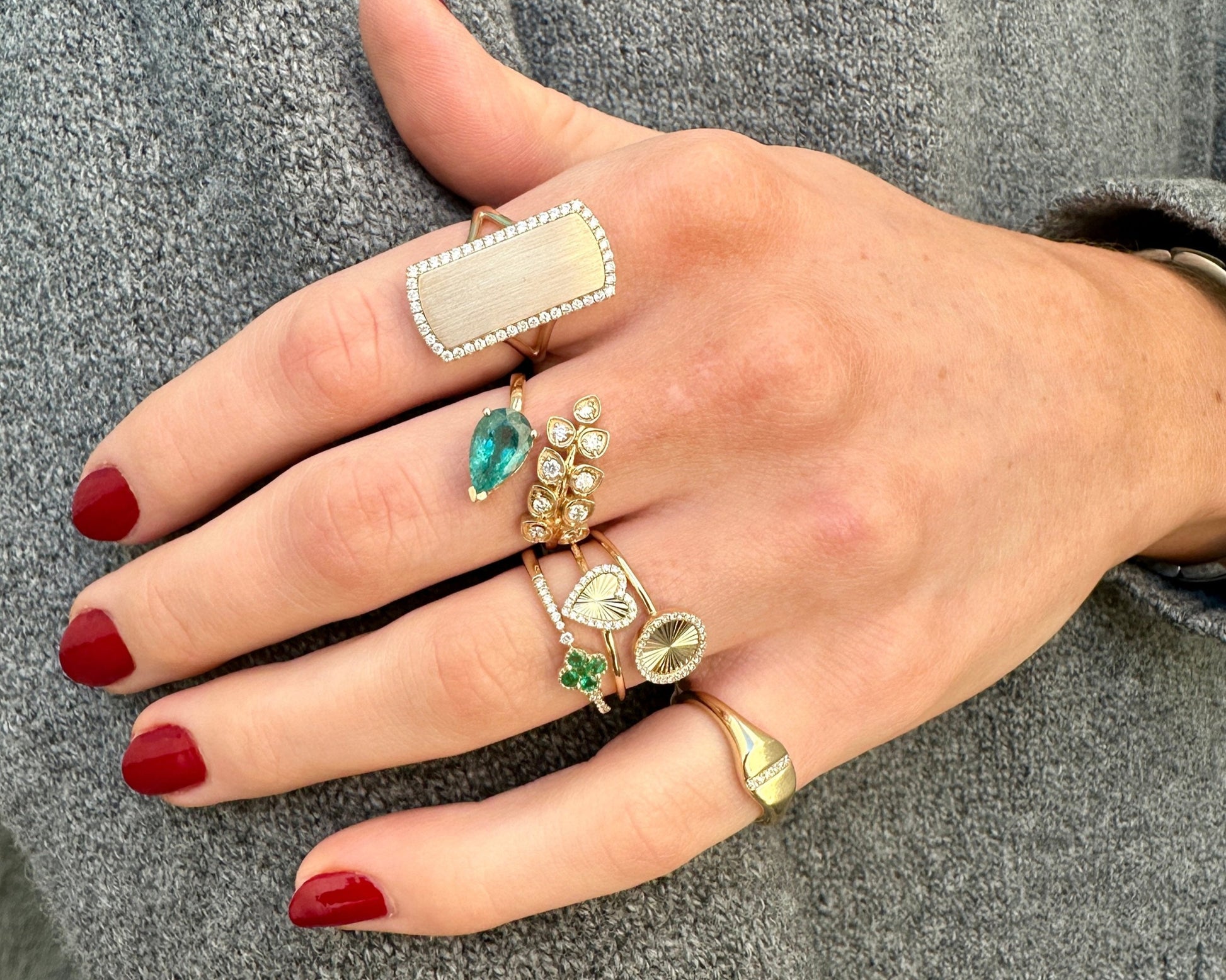 A hand with red nails showcases multiple rings, including the stunning LEE Gold Diamond Ring by AIRA, featuring a pear shape diamond in yellow gold, elegantly contrasted against a gray knitted sweater.