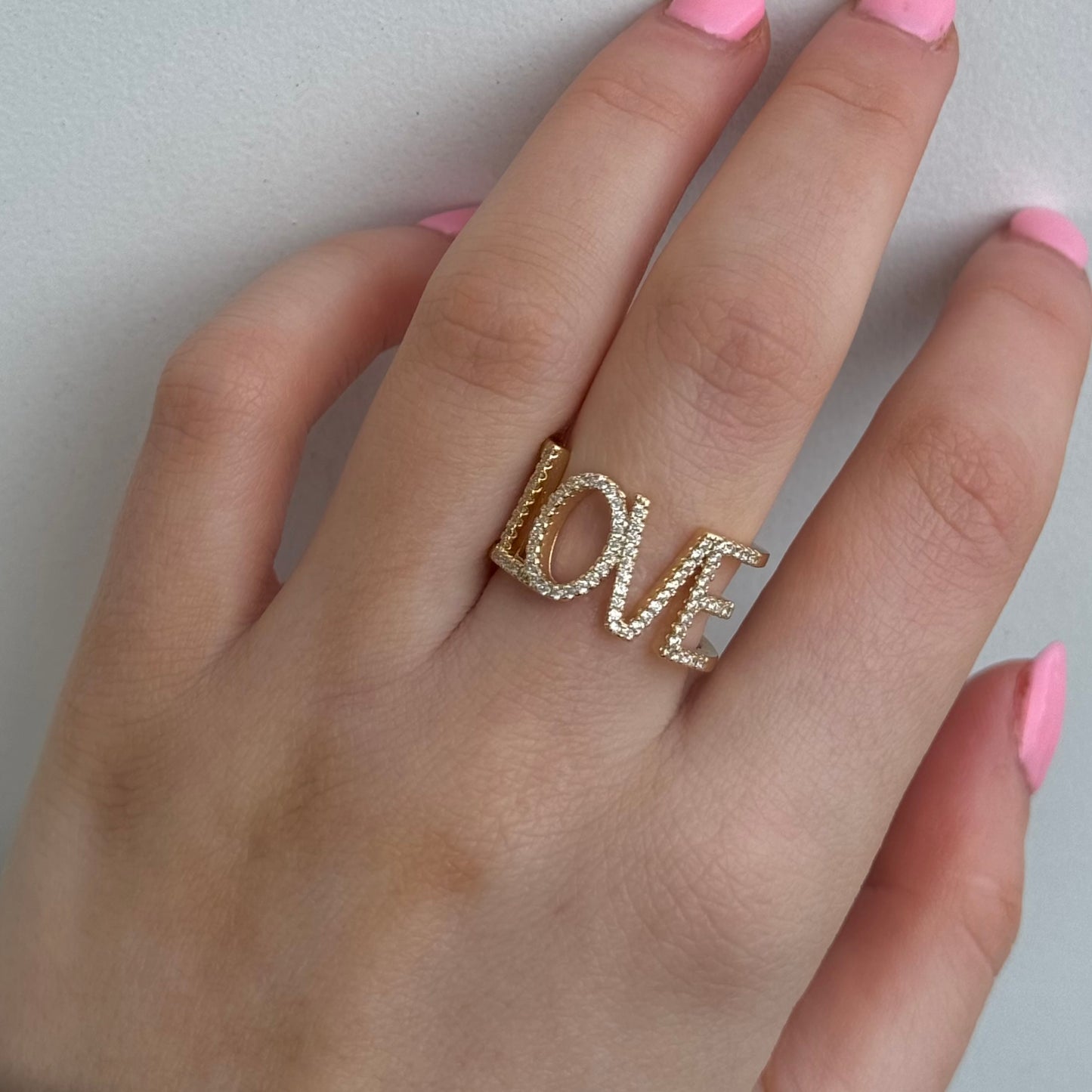 A hand with pink nails displays the Dylan James Jewelry LOVE Gold Diamond Ring, elegantly shimmering with a touch of cubic zirconia.