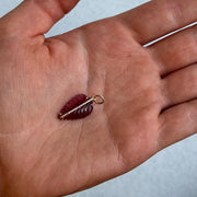 A hand holds the LEAF Charm by Dylan James Jewelry—a small, red, leaf-shaped pendant in 14kt yellow gold, featuring a gold loop and a row of tiny white stones along its center.
