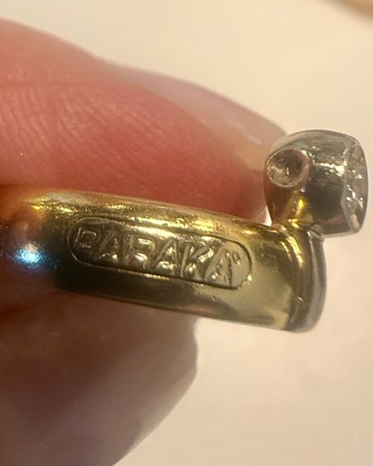 Gold ring with 'Bakka' branding held between fingers on a light background