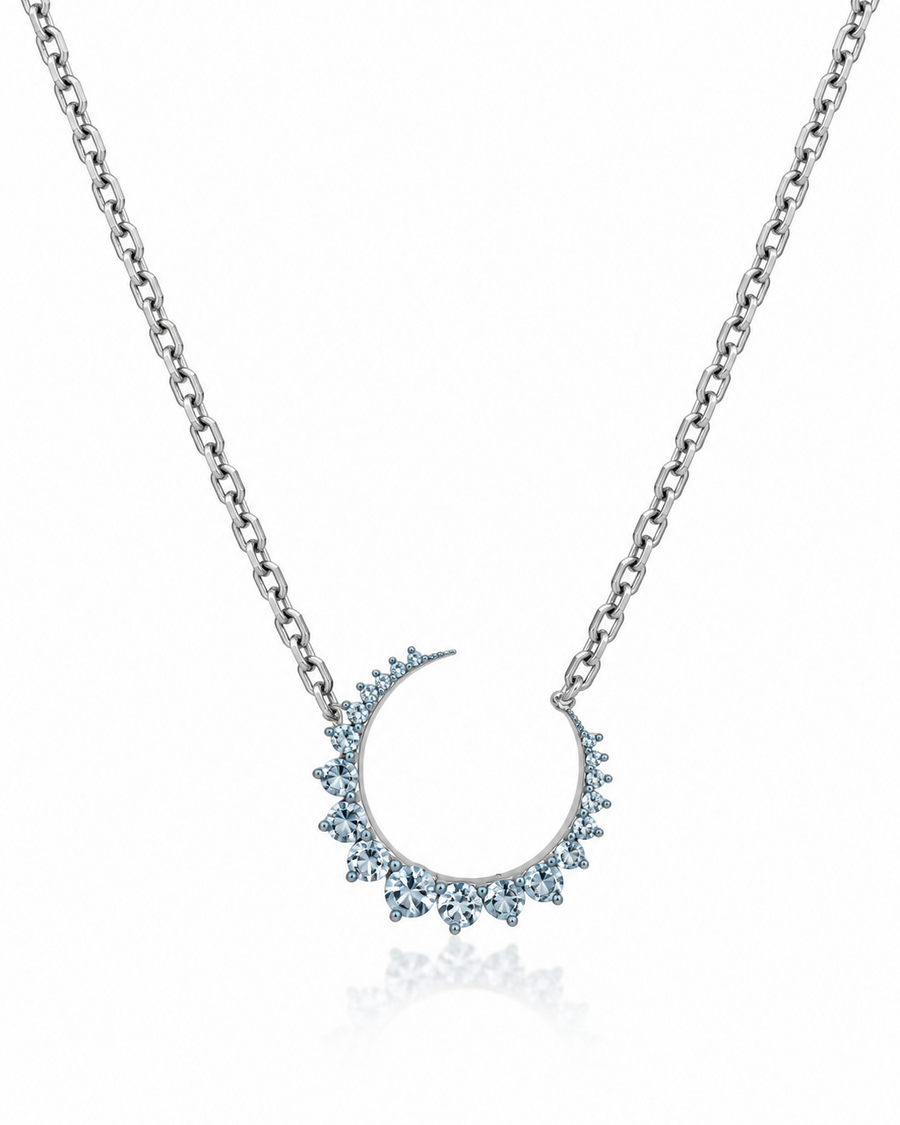 A silver-toned chain necklace featuring a crescent moon pendant adorned with small light blue gemstones, shown on a clean white background with a subtle reflection below.