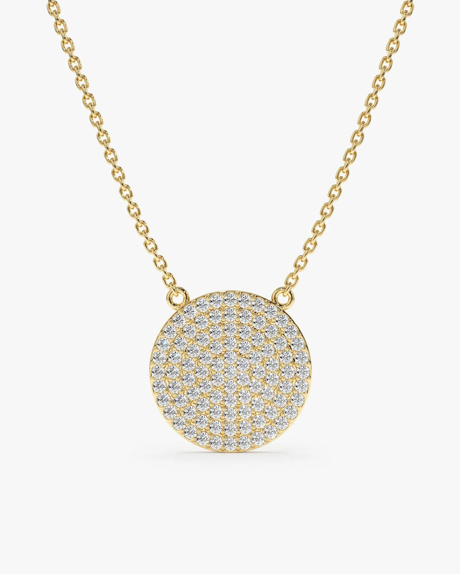 A gold chain necklace with a round, flat pendant paved with small clear stones, shown on a light gray background.