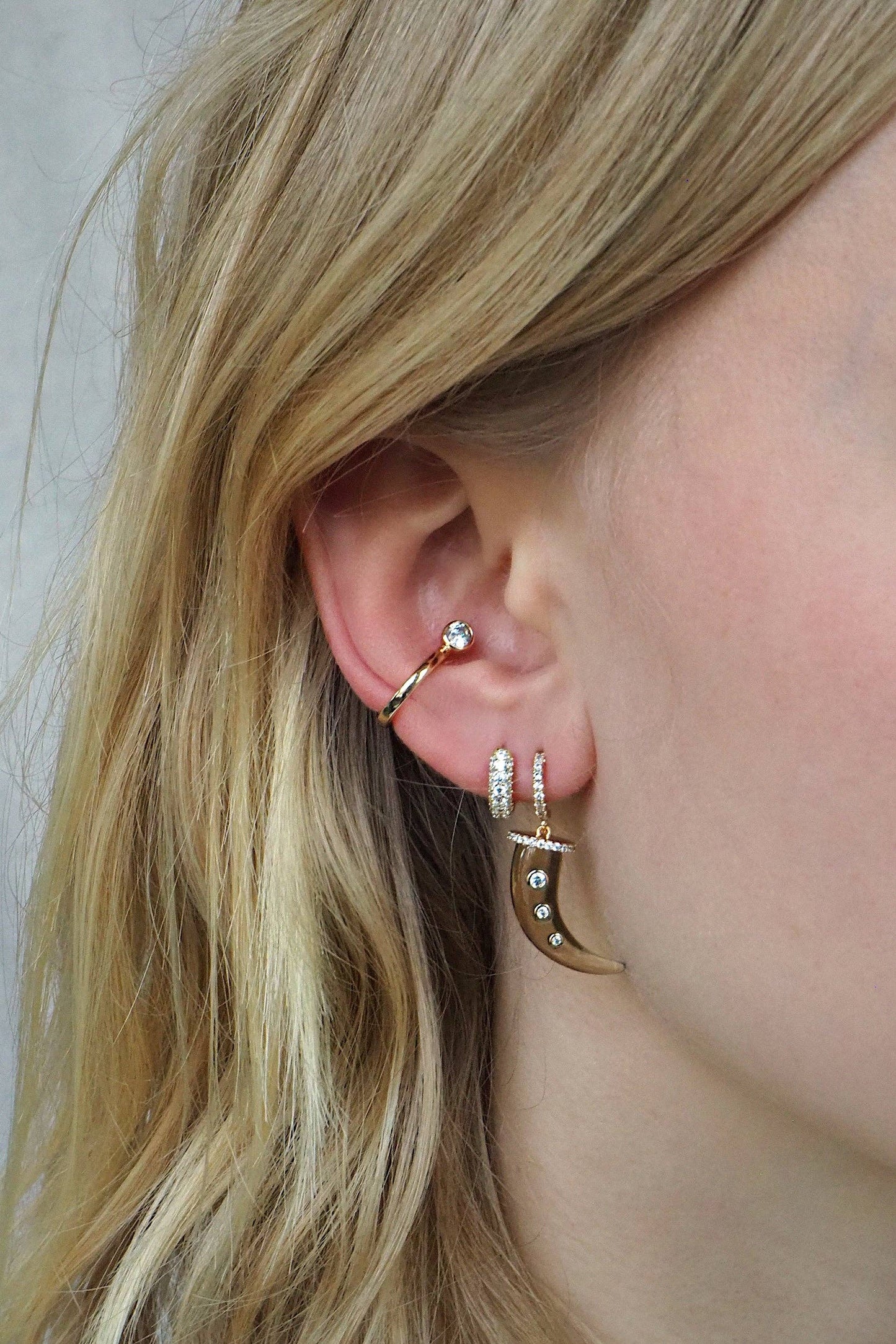 A close-up of a person's ear showcases an array of earrings, including the Lili Claspe REESE Conch Ear Cuff in 14kt gold plating and a small diamond stud. Two hoop earrings feature decorative elements with shimmering CZ stone accents, perfectly complementing the look. Partially visible is their flowing blonde hair.