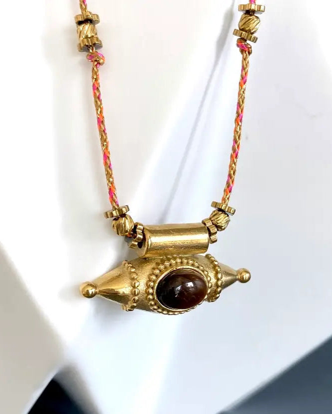 The Ma Cocotte Samara Amulet Necklace by Ma Cocotte features a gold-plated pendant with a central brown oval stone on a pink and yellow braided cord, shown against a white background.