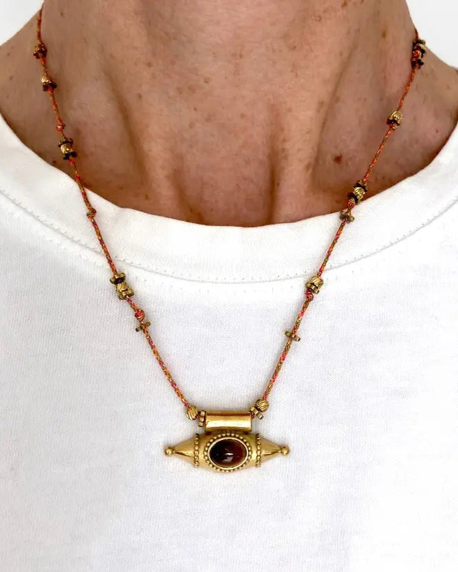 A person wearing a white shirt and the Ma Cocotte Samara Amulet Necklace by Ma Cocotte, featuring a gold-plated pendant with red stone on a beaded cord, styled as a protective amulet.