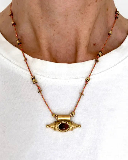A person wearing a white shirt and the Ma Cocotte Samara Amulet Necklace by Ma Cocotte, featuring a gold-plated pendant with red stone on a beaded cord, styled as a protective amulet.