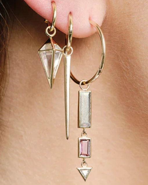 Close-up of an ear adorned with three geometric 9ct yellow gold earrings: a crystal pendant, a spike, and the SKINNY Point Plaque from Métier by Tomfoolery featuring a dangling bar with two colored gems and a triangle.